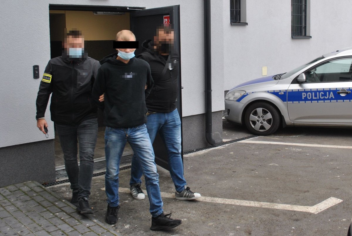 nieumundurowani policjanci prowadzą podejrzanego, w tle ściana budynku i radiowóz