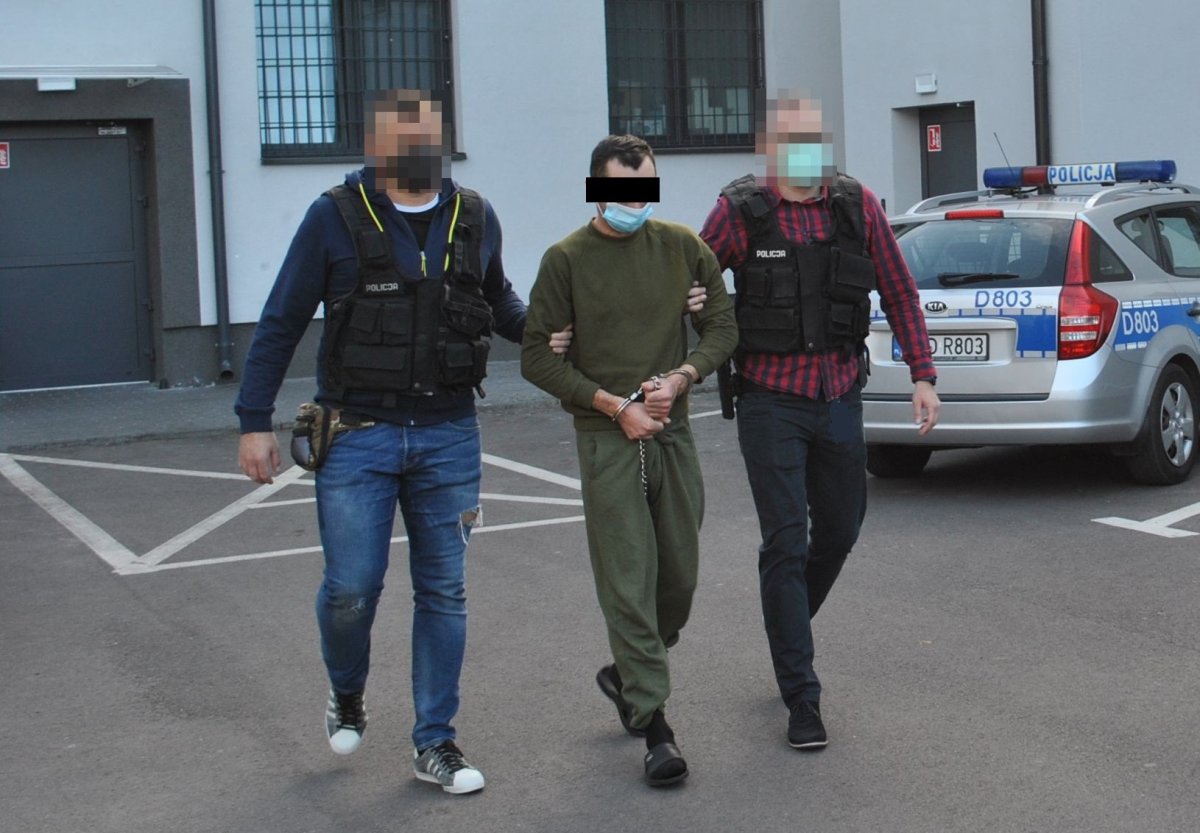 nieumundurowani policjanci prowadzą podejrzanego w kajdankach zespolonych, w tle ściana budynkku oraz radiowóz