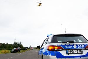 radiowóz policyjny, w tle samochody audi na miejscu wypadku, samochód straży pożarnej i śmigłowiec LPR