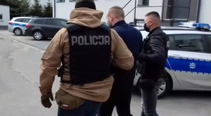 nieumundurowani policjanci prowadzą podejrzanego, w tle radiowóz policyjny