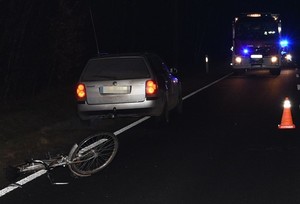 leżący na jezdni uszkodzony rower i stojący w pobliżu samochód marki Volkswagen Passat, w tle samochody Straży Pożarnej i Policji