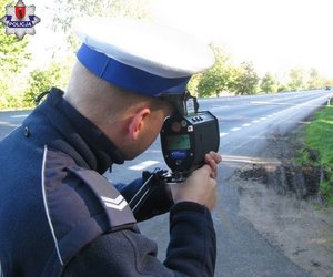 policjant z radarowym miernikiem prędkości, w tle asfaltowa jezdnia i przydrożne drzewa