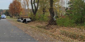 leżący na dachu w rowie uszkodzony samochód osobowy, w tle radiowóz oraz przydrożne drzewa