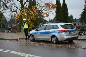 stojący na drodze radiowóz, obok policjant ruchu drogowego, w tle teren cmentarza