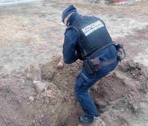 policjant stoi przy leżącym na gruntowym podłożu pocisku artyleryjskim