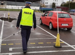 idący po placu manewrowym policjant, obok czerwony samochód fiat punto