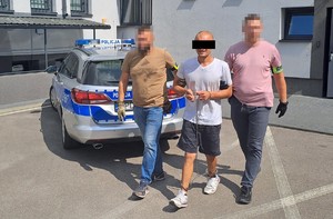 nieumundurowani policjanci prowadzą podejrzanego, w tle radiowóz i ściana budynku