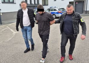 nieumundurowani policjanci prowadzą podejrzanego w kajdankach, w tle radiowozy i ściana budynku