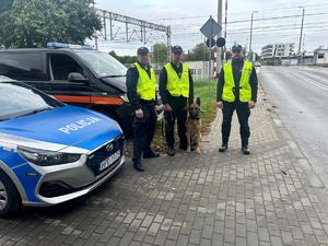 policjant i funkcjonariusze Straży Ochrony Kolei z psem służbowym, obok samochody służbowe, w tle tory kolejowe