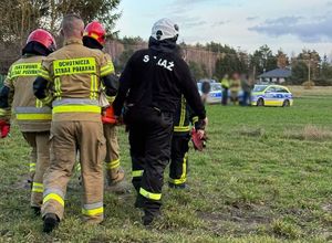 strażacy i druhowie ochotnicy niosą nosze z odnalezioną kobietą, w tle radiowozy