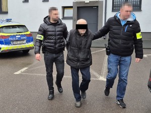 nieumundurowani policjanci prowadzą podejrzanego, w tle radiowóz i ściana budynku