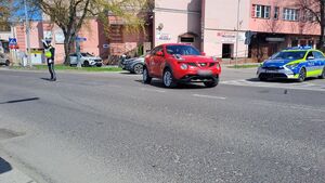 regulujący ruchem na miejscu wypadku policjant z drogówki, stojący na skrzyżowaniu czerwony samochód marki Nissan, stojący na skrzyżowaniu radiowóz, w tle budynki