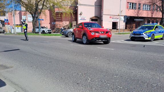 regulujący ruchem na miejscu wypadku policjant z drogówki, stojący na skrzyżowaniu czerwony samochód marki Nissan, stojący na skrzyżowaniu radiowóz, w tle budynki