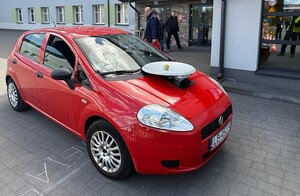 czerwony samochód osobowy z talerzem na pokrywie silnika, w tle budynek i policjant